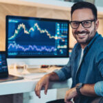 a smiling investor sitting in front of two computer screens which are displaying stock or cryptocurr