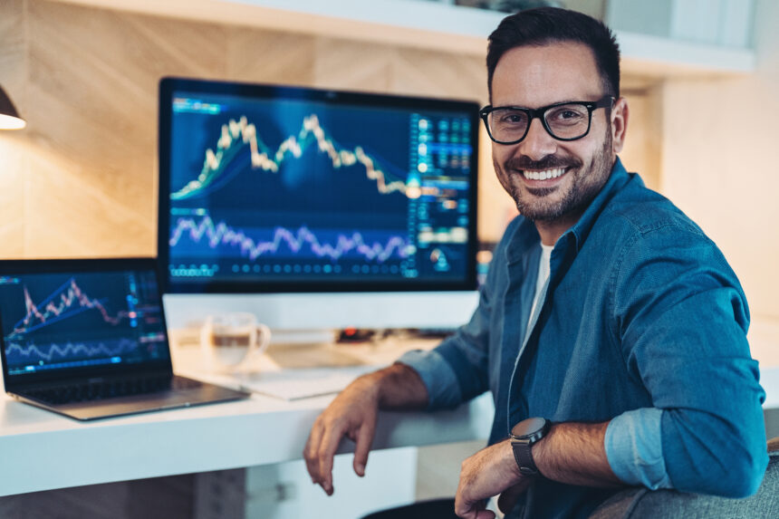 a smiling investor sitting in front of two computer screens which are displaying stock or cryptocurr