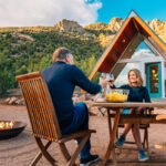 two people smile while clinking glasses in an outdoor setting by an a frame house and a fire pit