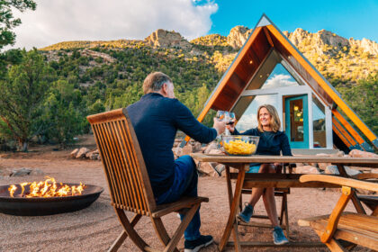 two people smile while clinking glasses in an outdoor setting by an a frame house and a fire pit